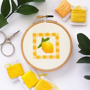 May include: A cross-stitch embroidery hoop featuring a yellow lemon design within a gingham border. The embroidery is set in a wooden hoop, surrounded by yellow thread spools, scissors, and greenery. The color palette is bright and cheerful.