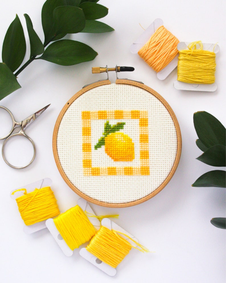 May include: A cross-stitch embroidery hoop featuring a yellow lemon design within a gingham border. The embroidery is set in a wooden hoop, surrounded by yellow thread spools, scissors, and greenery. The color palette is bright and cheerful.