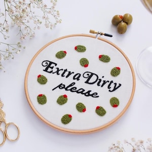 May include: Embroidered hoop art with the text "Extra Dirty please" in black cursive font. The hoop is filled with green and red embroidered olives.