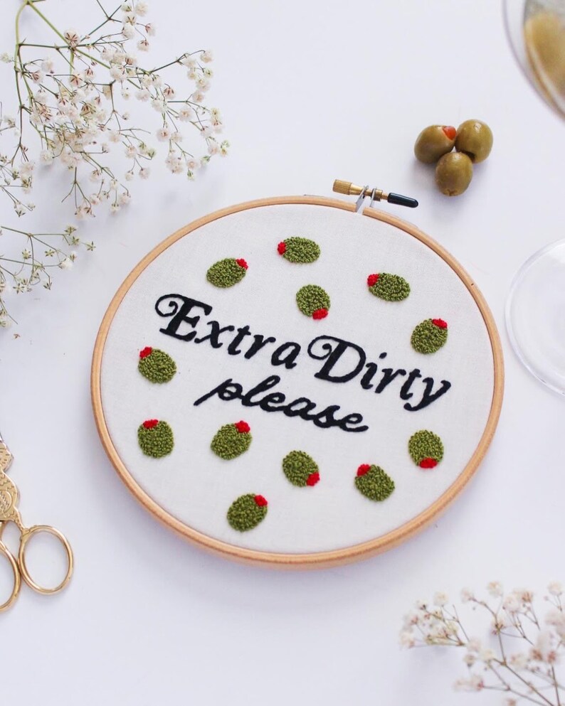 May include: Embroidered hoop art with the text "Extra Dirty please" in black cursive font. The hoop is filled with green and red embroidered olives.