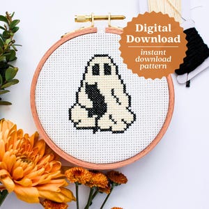 May include: A cross stitch pattern of a ghost sitting down, framed in a wooden hoop. The hoop is on a white surface with orange flowers.