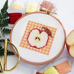 Puede incluir: Un aro de bordado de punto de cruz con un dise&ntilde;o de manzana roja sobre un fondo melocot&oacute;n. El aro est&aacute; rodeado de carretes de hilo de bordar en varios tonos de rojo, naranja y amarillo.