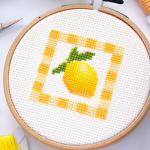 May include: A cross-stitch embroidery hoop featuring a yellow lemon design with green leaves inside a yellow and orange checkered border. The embroidery is set in a wooden hoop, with yellow thread and scissors visible.