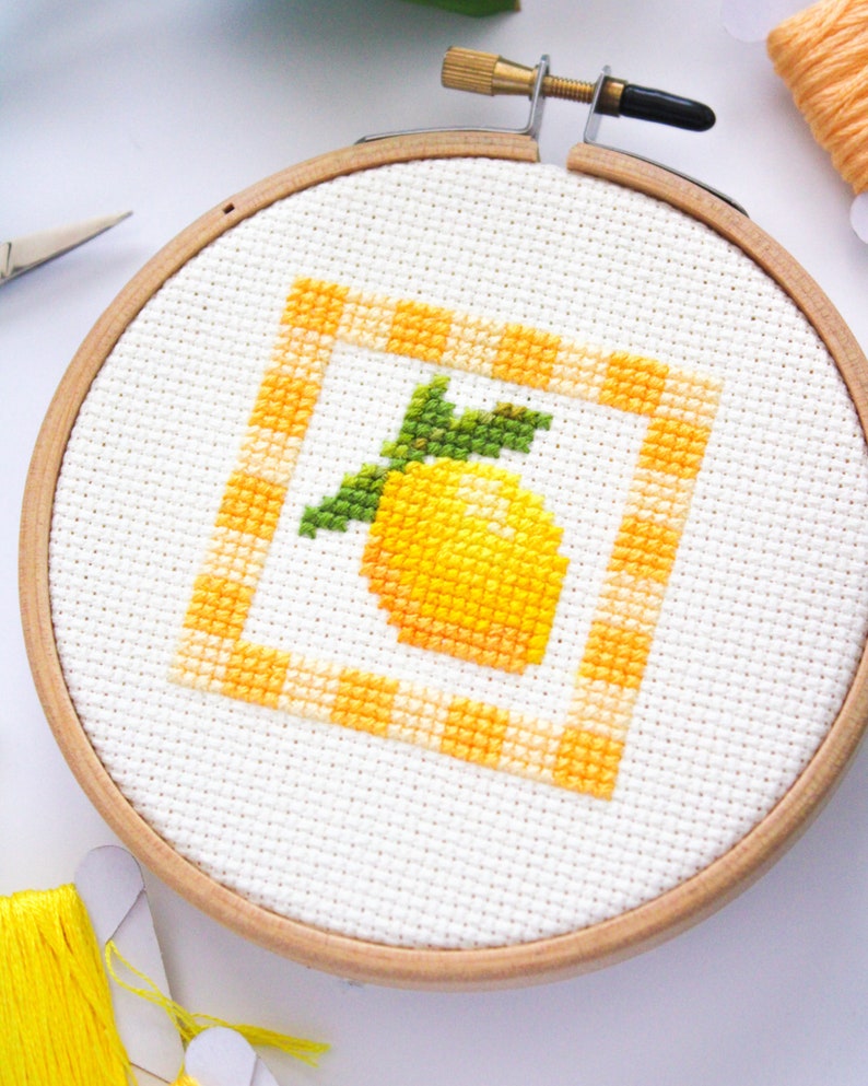 May include: A cross-stitch embroidery hoop featuring a yellow lemon design with green leaves inside a yellow and orange checkered border. The embroidery is set in a wooden hoop, with yellow thread and scissors visible.