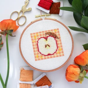 Puede incluir: Primer plano de un proyecto de bordado de punto de cruz terminado de una manzana sobre un fondo de cuadros. El bordado est&aacute; enmarcado en un aro de madera. El aro est&aacute; rodeado de carretes de hilo de bordar naranja y rojo y un par de tijeras doradas.