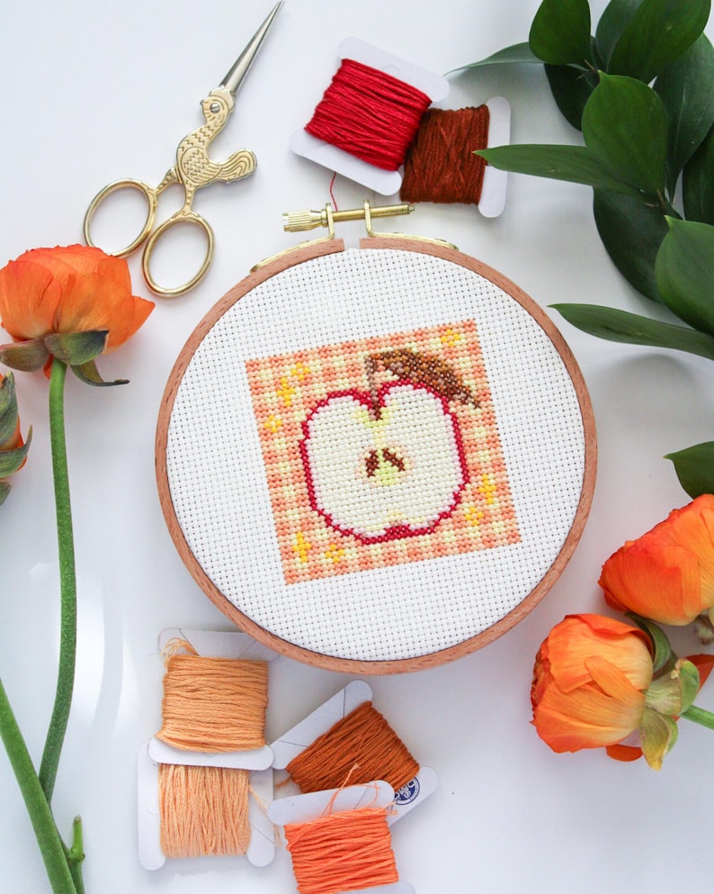 Puede incluir: Primer plano de un proyecto de bordado de punto de cruz terminado de una manzana sobre un fondo de cuadros. El bordado est&aacute; enmarcado en un aro de madera. El aro est&aacute; rodeado de carretes de hilo de bordar naranja y rojo y un par de tijeras doradas.