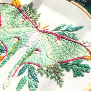 May include: Embroidered hoop art featuring a green luna moth with pink and yellow accents surrounded by green leaves.