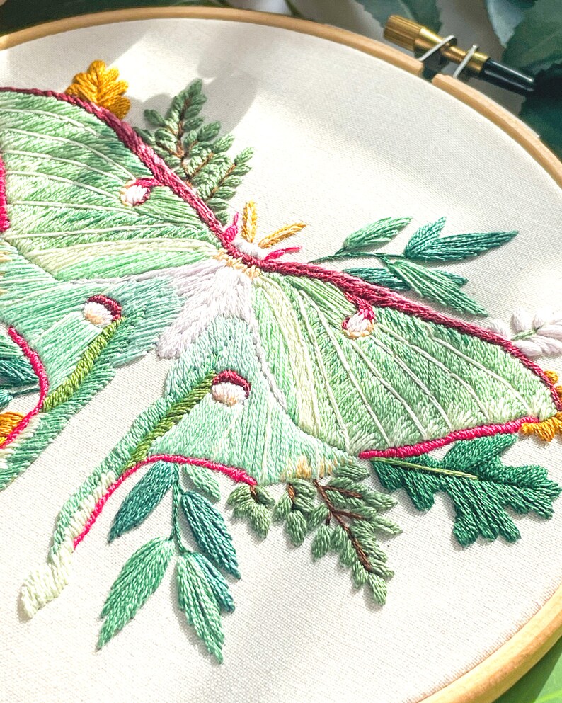 May include: Embroidered hoop art featuring a green luna moth with pink and yellow accents surrounded by green leaves.