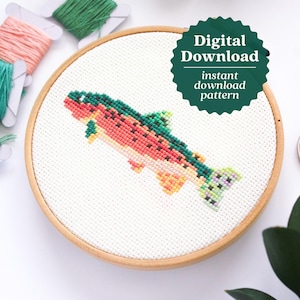 May include: A finished cross-stitch embroidery project in a wooden hoop, depicting a colorful trout fish. The hoop is on a white surface with green leaves and pink and green embroidery floss.