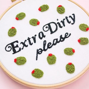 May include: Embroidered hoop art with the text "Extra Dirty please" surrounded by green olives with red centers.