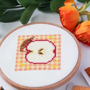 Puede incluir: Un aro de bordado de punto de cruz con un dise&ntilde;o de manzana roja. La manzana est&aacute; cortada por la mitad y el interior es blanco con semillas marrones. El fondo es un patr&oacute;n de cuadros de color melocot&oacute;n y naranja.