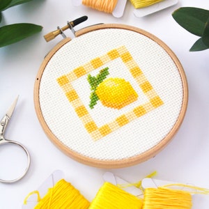 May include: A cross-stitch embroidery hoop featuring a yellow lemon design with green leaves and a yellow and white checkered border. The embroidery is set in a wooden hoop, surrounded by yellow and orange embroidery floss and a pair of scissors.