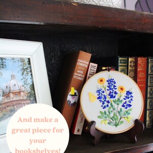 Puede incluir: Un aro de bordado blanco con un dise&ntilde;o de flores de campanilla azul, flores amarillas y una mariposa. El aro est&aacute; en un soporte de madera y se exhibe en una estanter&iacute;a con libros.