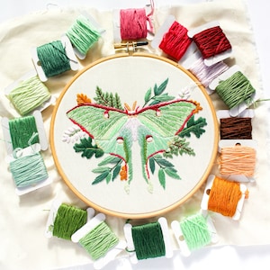 May include: A close-up of an embroidery hoop with a green and white luna moth embroidered on a white fabric. The hoop is surrounded by spools of thread in various colors, including green, red, and brown.