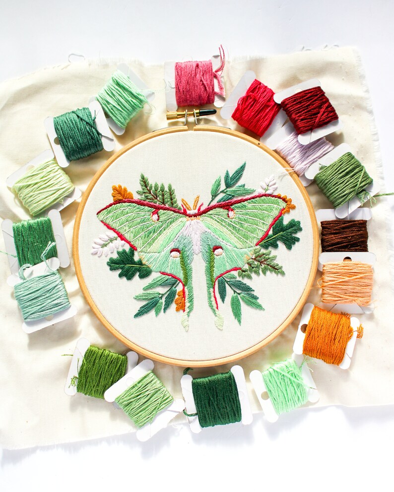 May include: A close-up of an embroidery hoop with a green and white luna moth embroidered on a white fabric. The hoop is surrounded by spools of thread in various colors, including green, red, and brown.