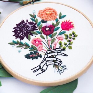 Puede incluir: Arte de bordado en aro que presenta una mano de esqueleto sosteniendo un ramo de flores coloridas. Las flores son rosas, rojas, moradas, naranjas y verdes.
