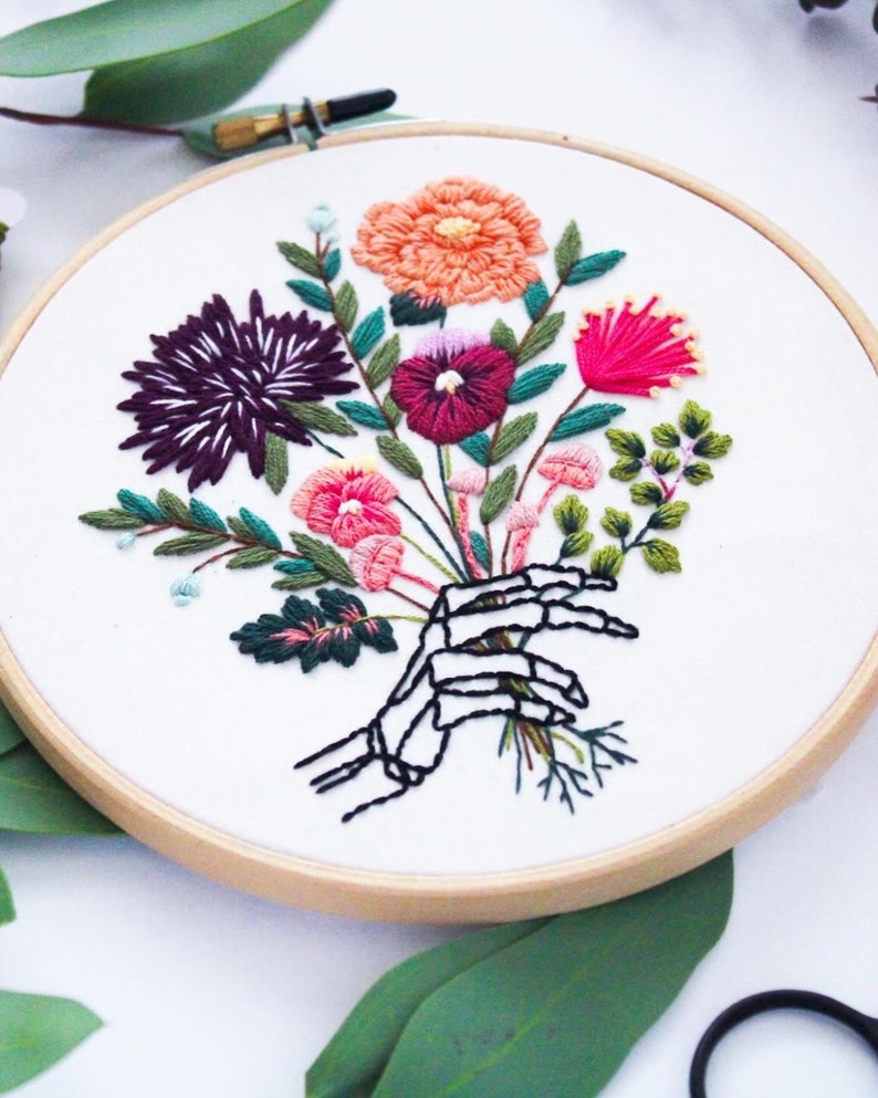 Puede incluir: Arte de bordado en aro que presenta una mano de esqueleto sosteniendo un ramo de flores coloridas. Las flores son rosas, rojas, moradas, naranjas y verdes.