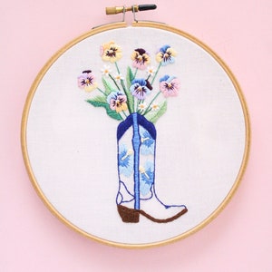 May include: Embroidered hoop art featuring a white cowboy boot with blue and white floral designs, filled with a bouquet of colorful pansies.