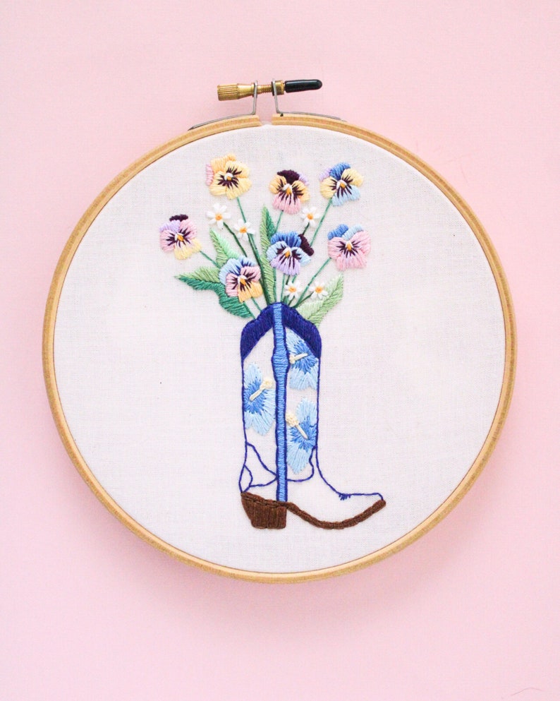 May include: Embroidered hoop art featuring a white cowboy boot with blue and white floral designs, filled with a bouquet of colorful pansies.