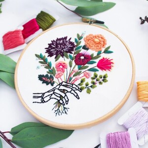 Puede incluir: Arte de bordado en aro que presenta una mano de esqueleto sosteniendo un ramo de flores coloridas. Las flores son rosas, rojas, naranjas, moradas y verdes. El aro est&aacute; hecho de madera y tiene un fondo de tela blanca.