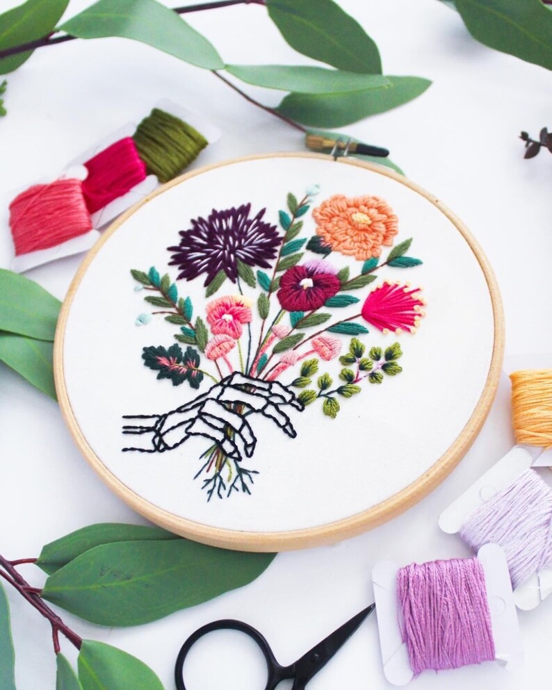 Puede incluir: Arte de bordado en aro que presenta una mano de esqueleto sosteniendo un ramo de flores coloridas. Las flores son rosas, rojas, naranjas, moradas y verdes. El aro est&aacute; hecho de madera y tiene un fondo de tela blanca.