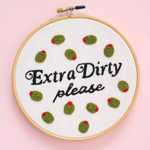 May include: Embroidered hoop art with the text "Extra Dirty Please" in black cursive font. The hoop is filled with green olives with red centers.
