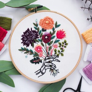 Puede incluir: Arte de bordado en aro que presenta una mano de esqueleto sosteniendo un ramo de flores coloridas. Las flores son en tonos de rosa, rojo, morado, naranja y verde. El fondo es blanco.