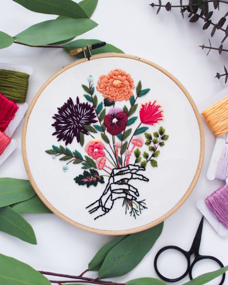 Puede incluir: Arte de bordado en aro que presenta una mano de esqueleto sosteniendo un ramo de flores coloridas. Las flores son en tonos de rosa, rojo, morado, naranja y verde. El fondo es blanco.