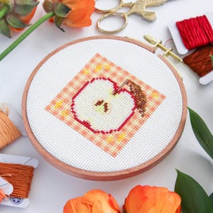 Puede incluir: Primer plano de un proyecto de bordado de punto de cruz terminado en un aro de madera. El dise&ntilde;o presenta una manzana roja con un centro blanco y un tallo marr&oacute;n sobre un fondo a cuadros. El bordado est&aacute; rodeado de flores naranjas e hilo de bordar.
