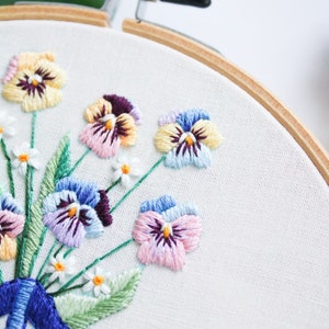 May include: Close-up of an embroidery hoop with a floral design. The design features a bouquet of purple, pink, yellow, and blue pansies with white accents. The pansies are embroidered on white fabric.