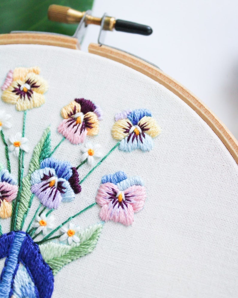 May include: Close-up of an embroidery hoop with a floral design. The design features a bouquet of purple, pink, yellow, and blue pansies with white accents. The pansies are embroidered on white fabric.