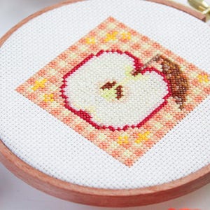 Puede incluir: Primer plano de un aro de bordado de punto de cruz con un dise&ntilde;o de manzana roja. La manzana es blanca con un contorno rojo y un tallo marr&oacute;n. El fondo es un patr&oacute;n de cuadros de color melocot&oacute;n y naranja.