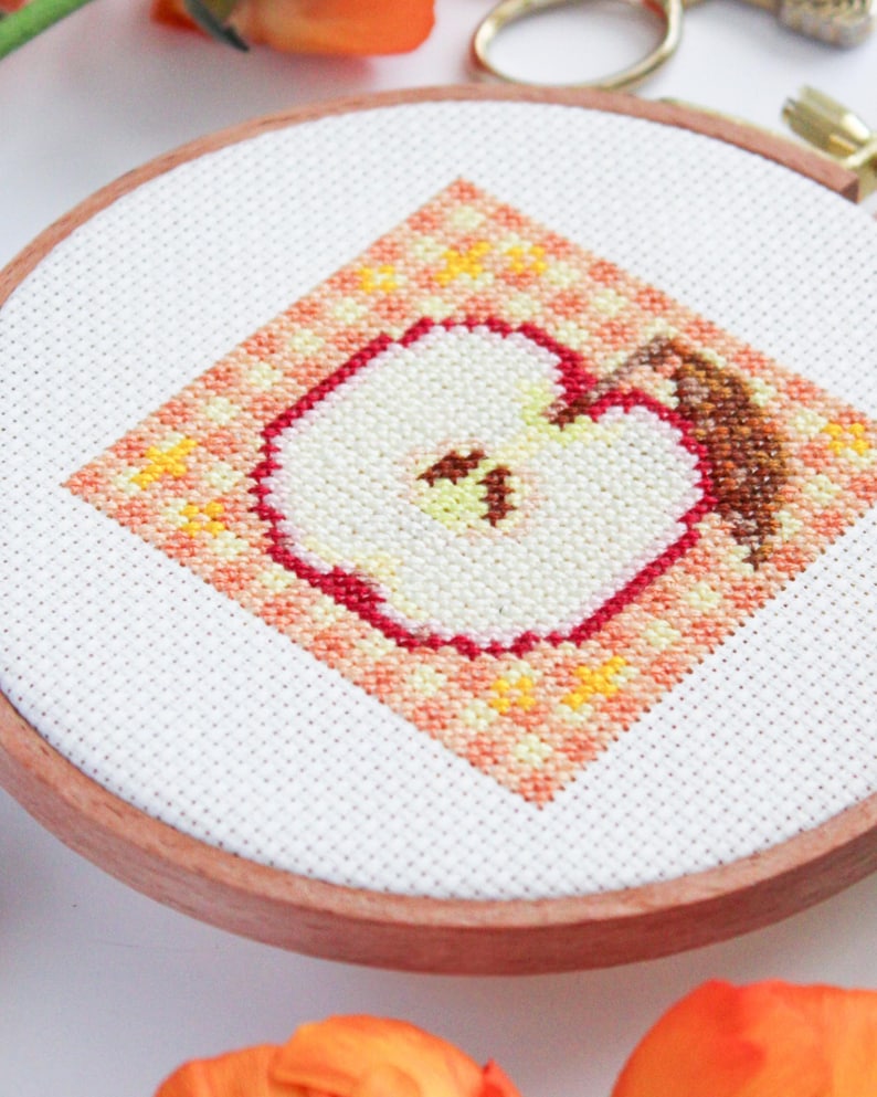 Puede incluir: Primer plano de un aro de bordado de punto de cruz con un dise&ntilde;o de manzana roja. La manzana es blanca con un contorno rojo y un tallo marr&oacute;n. El fondo es un patr&oacute;n de cuadros de color melocot&oacute;n y naranja.