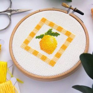 May include: A cross-stitch embroidery hoop featuring a yellow lemon design with green leaves inside an orange and white checkered square. The embroidery is set in a wooden hoop, with yellow thread and scissors nearby.