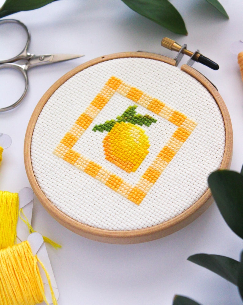 May include: A cross-stitch embroidery hoop featuring a yellow lemon design with green leaves inside an orange and white checkered square. The embroidery is set in a wooden hoop, with yellow thread and scissors nearby.