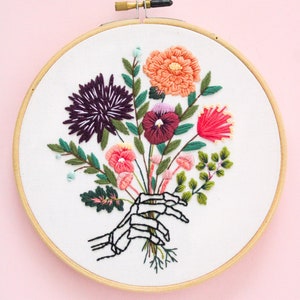 Puede incluir: Arte de bordado en aro que presenta una mano de esqueleto sosteniendo un ramo de flores coloridas, incluyendo rosa, rojo, naranja, morado y verde.