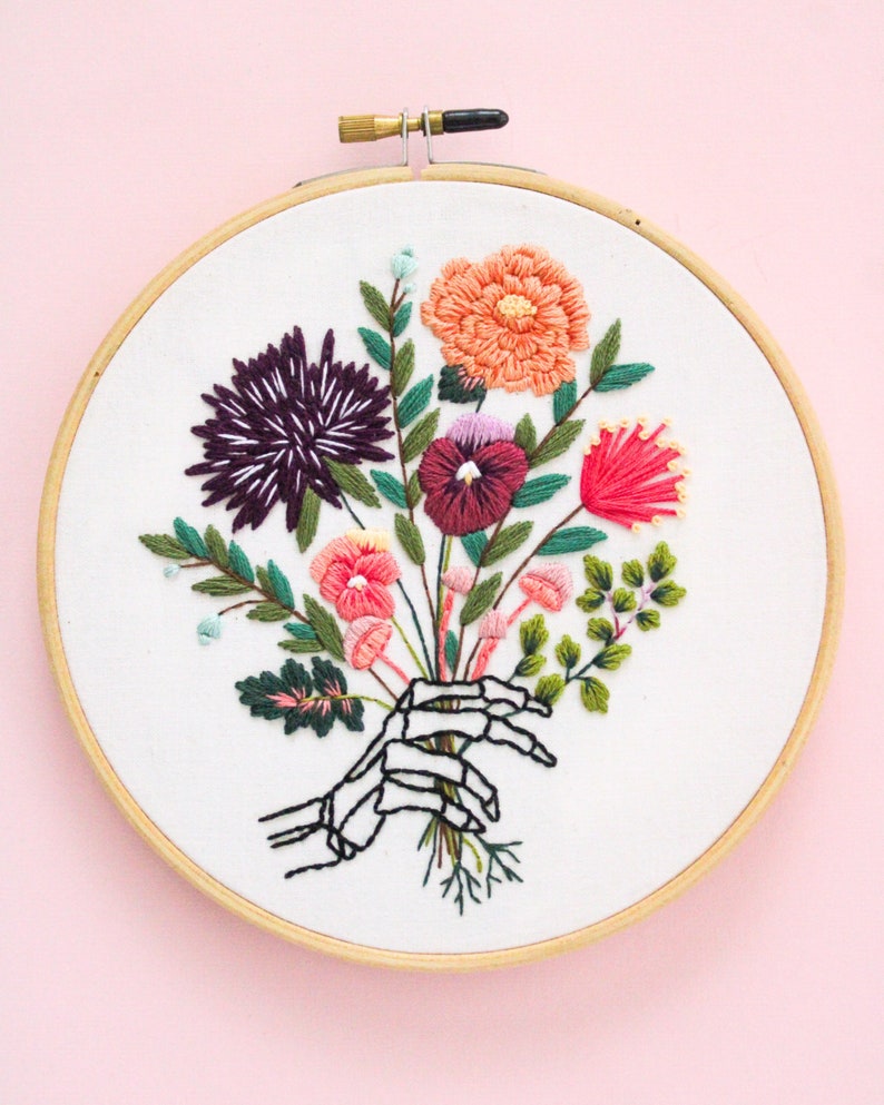 Puede incluir: Arte de bordado en aro que presenta una mano de esqueleto sosteniendo un ramo de flores coloridas, incluyendo rosa, rojo, naranja, morado y verde.