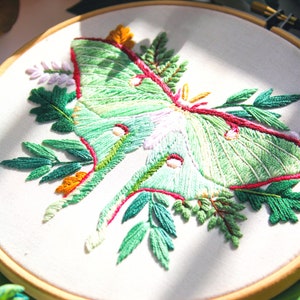 May include: Embroidered hoop art featuring a green and pink Luna moth surrounded by green leaves and flowers. The hoop is made of wood and has a white fabric background.