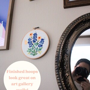 Puede incluir: Un aro de bordado blanco con un dise&ntilde;o de flores de campanilla azul y una mariposa amarilla. El aro est&aacute; colgado en una pared con otras obras de arte enmarcadas. El texto "Finished hoops look great on art gallery walls!" est&aacute; escrito en un c&iacute;rculo blanco en la pared.