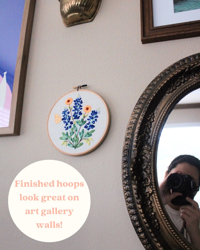 Puede incluir: Un aro de bordado blanco con un dise&ntilde;o de flores de campanilla azul y una mariposa amarilla. El aro est&aacute; colgado en una pared con otras obras de arte enmarcadas. El texto "Finished hoops look great on art gallery walls!" est&aacute; escrito en un c&iacute;rculo blanco en la pared.