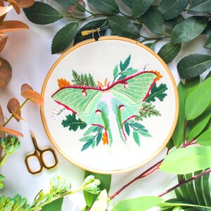 May include: Embroidered hoop art featuring a green luna moth with pink and red accents surrounded by green leaves and flowers.