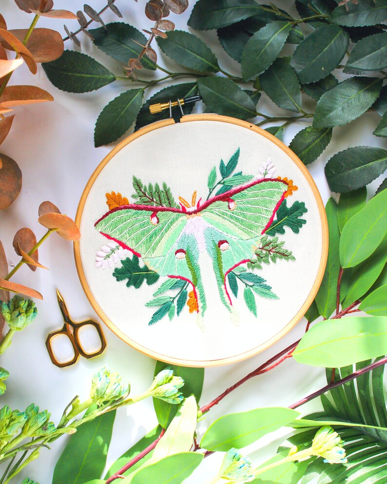 May include: Embroidered hoop art featuring a green luna moth with pink and red accents surrounded by green leaves and flowers.