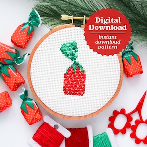 May include: A cross stitch pattern of a strawberry candy, with green leaves and red and white details. The pattern is displayed in a wooden embroidery hoop, with red and green embroidery floss and red scissors nearby. The text "Digital Download instant download pattern" is on a red starburst.