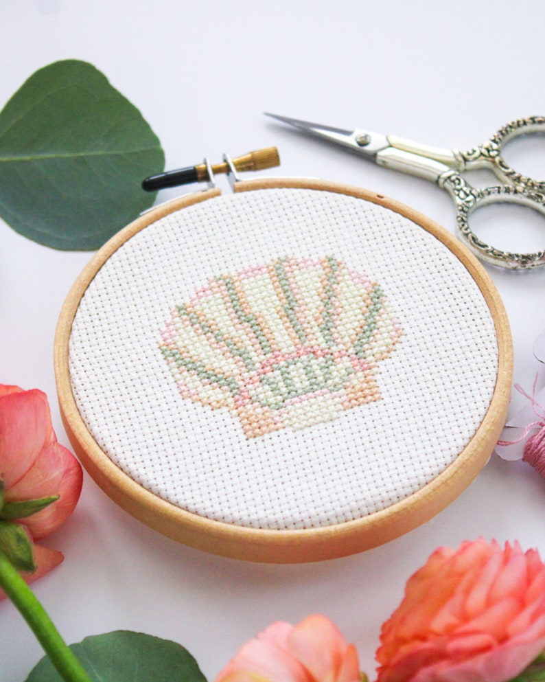 K&ouml;nnte beinhalten: Nahaufnahme eines fertigen Kreuzstich-Stickprojekts mit einer rosa und gr&uuml;nen Muschel in einem h&ouml;lzernen Stickrahmen.