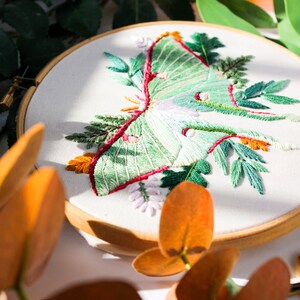 May include: Embroidered hoop art featuring a green luna moth with red and white accents surrounded by green foliage.