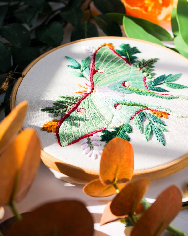 May include: Embroidered hoop art featuring a green luna moth with red and white accents surrounded by green foliage.