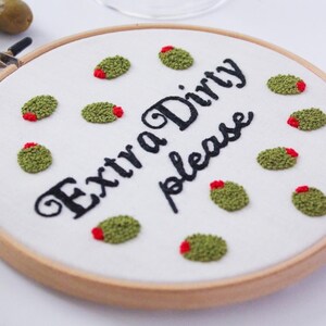 May include: Embroidered hoop with the text "Extra Dirty Please" in black cursive font. The hoop is decorated with green and red embroidered olives.