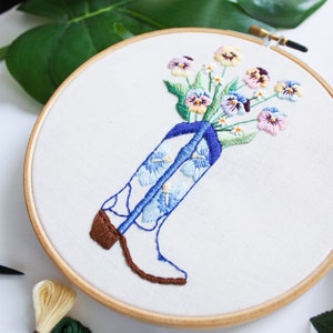 May include: Embroidered hoop art featuring a white cowboy boot with blue and white floral embroidery. A bouquet of colorful pansies are arranged in the boot's top.