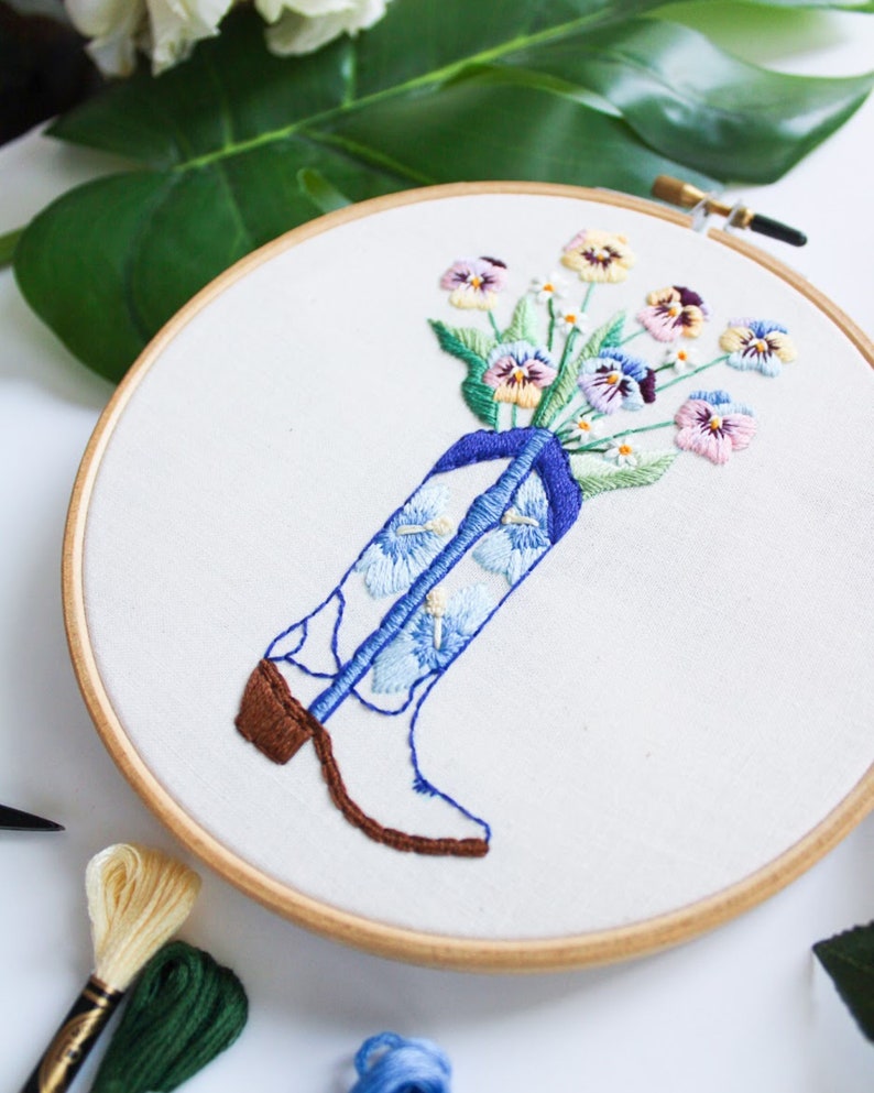 May include: Embroidered hoop art featuring a white cowboy boot with blue and white floral embroidery. A bouquet of colorful pansies are arranged in the boot's top.
