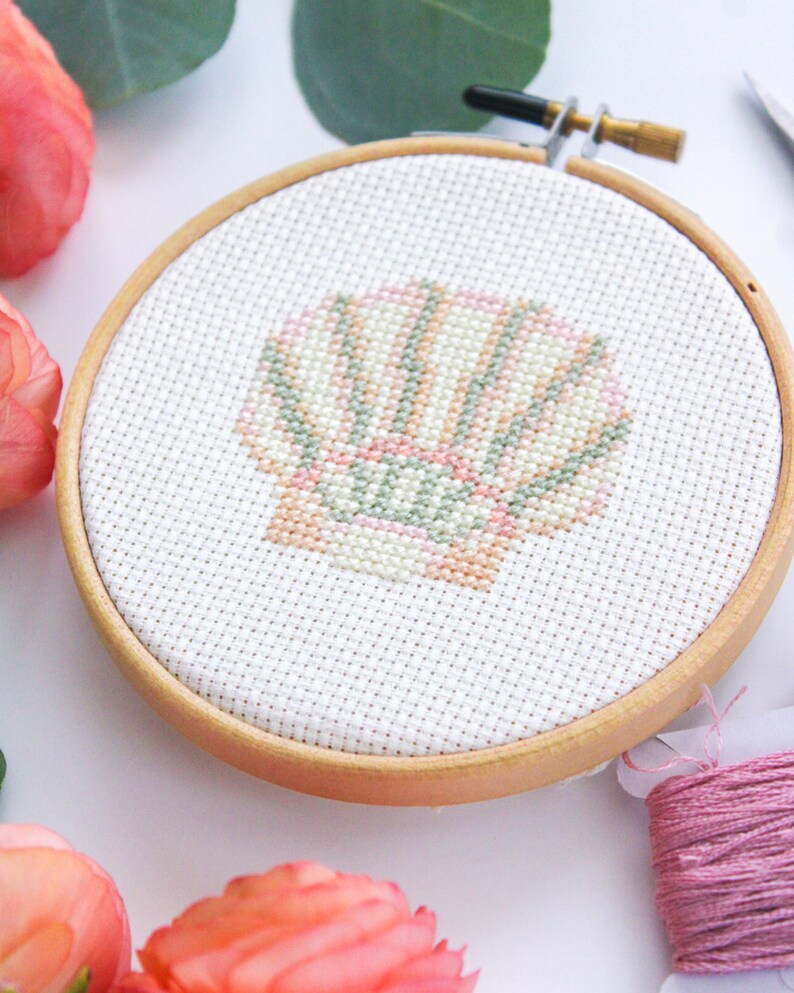 K&ouml;nnte beinhalten: Nahaufnahme eines fertigen Kreuzstich-Stickprojekts in einem Holzreifen. Das Design ist eine Muschel mit rosa, gr&uuml;nen und wei&szlig;en Streifen.